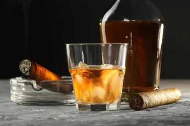Bottle, glass of whiskey with ice cubes and cigars on grey table, closeup