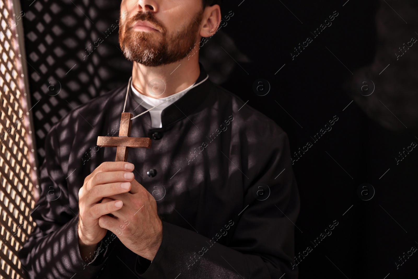 Photo of Catholic priest in cassock holding cross and praying to God in confessional, closeup
