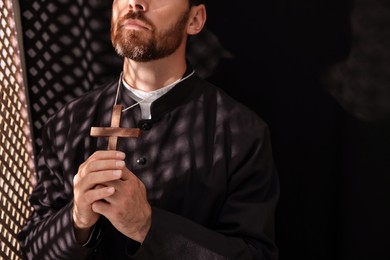 Photo of Catholic priest in cassock holding cross and praying to God in confessional, closeup