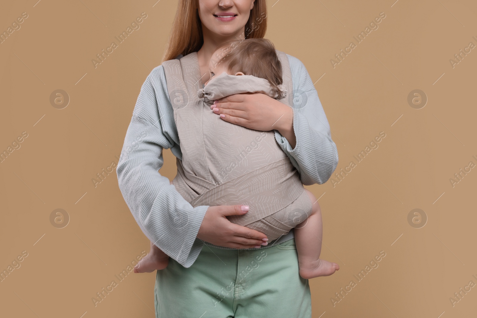 Photo of Mother holding her child in sling (baby carrier) on light brown background, closeup