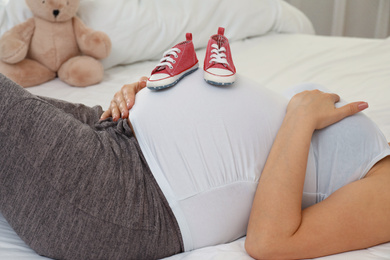 Photo of Pregnant woman with cute baby shoes on bed at home, closeup