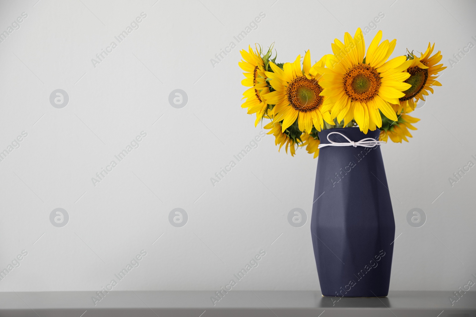 Photo of Bouquet of beautiful sunflowers in vase on table near light wall. Space for text