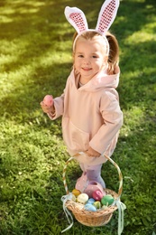 Cute little girl with bunny ears and basket of Easter eggs in park