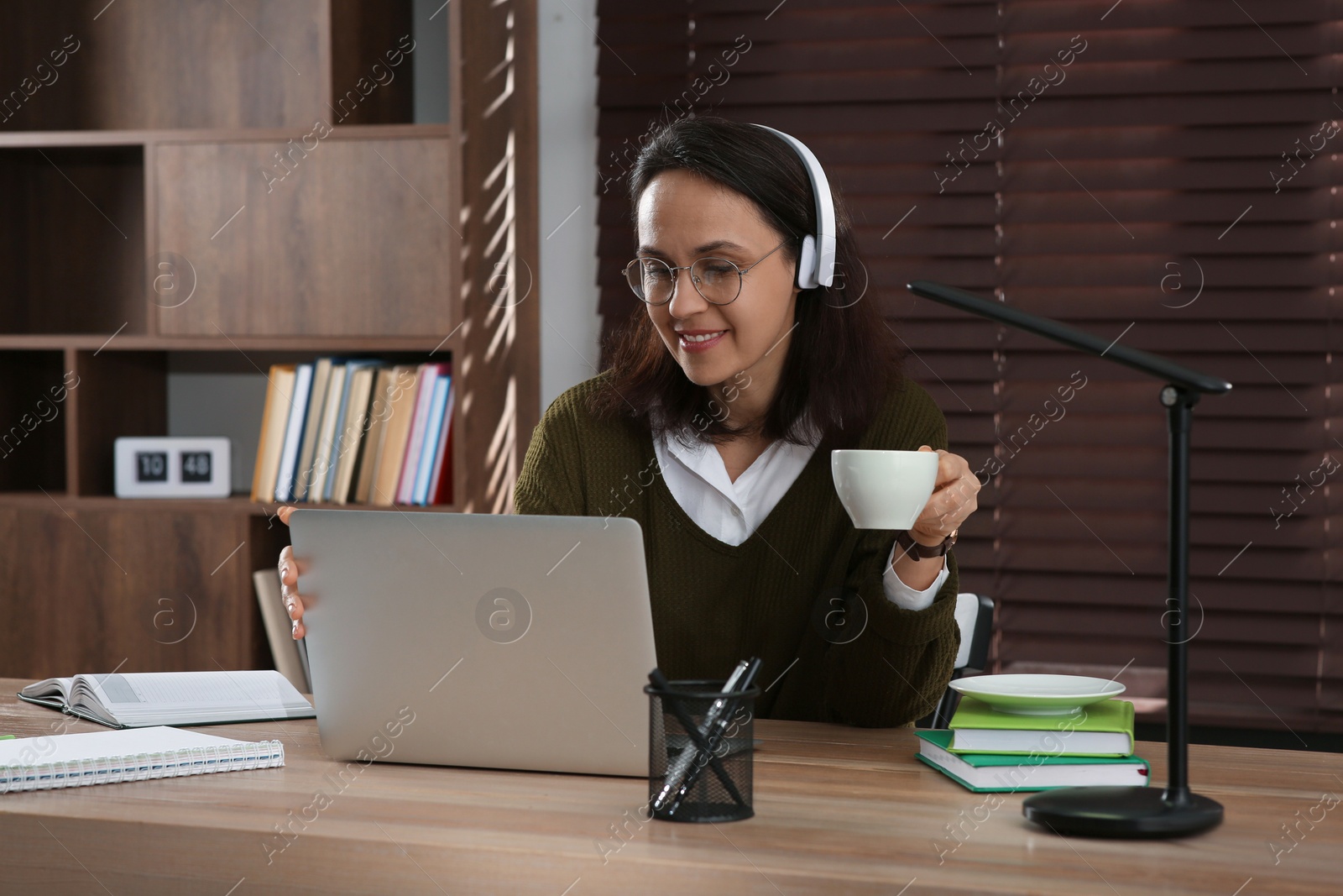 Photo of Woman with modern laptop and headphones drinking tea while learning at table indoors