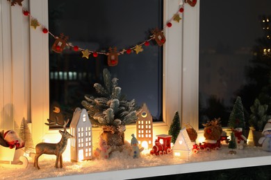 Photo of Christmas atmosphere. Beautiful glowing houses, artificial snow and toys on window sill indoors