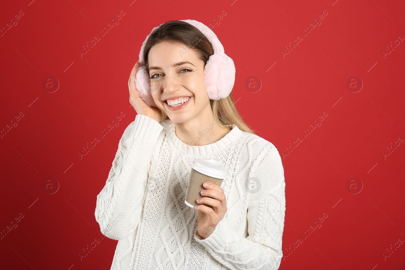 Photo of Happy woman with cup of drink wearing warm earmuffs on red background