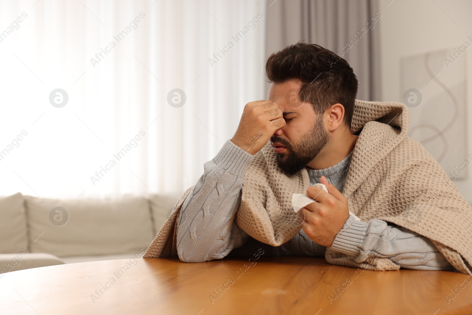 Photo of Sick man wrapped in blanket with tissue at wooden table indoors, space for text. Cold symptoms