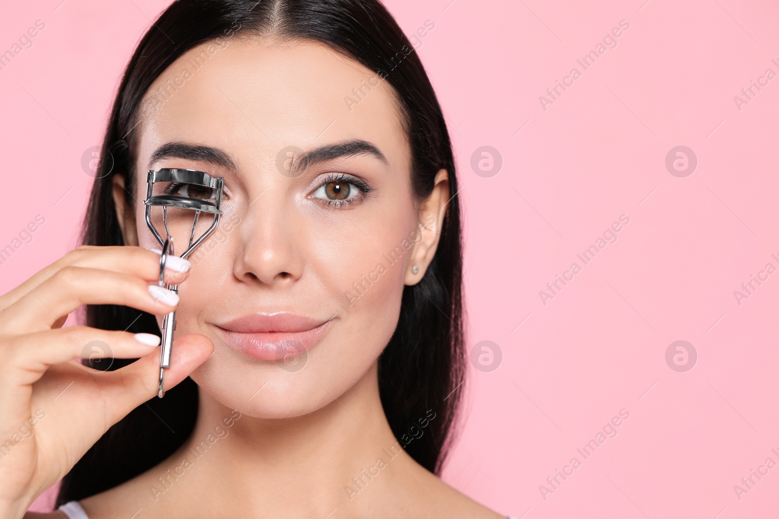 Photo of Beautiful young woman using eyelash curler on light pink background, space for text