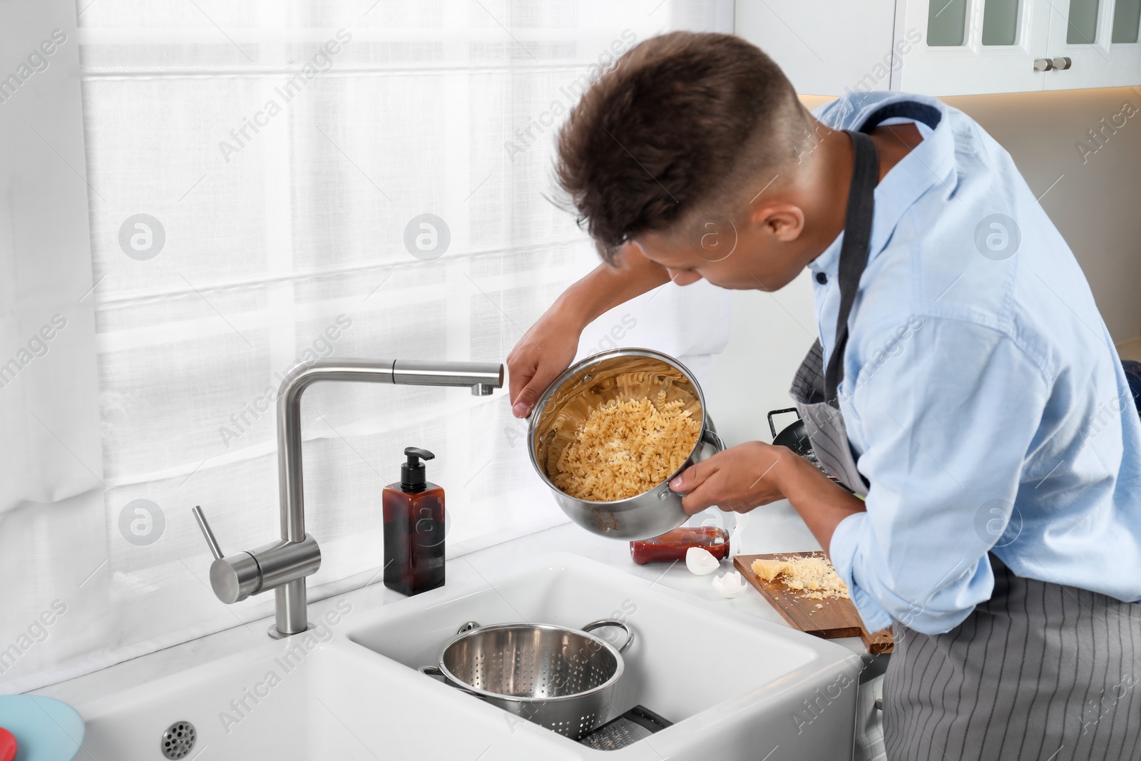 Photo of Man draining water from saucepan with pasta in messy kitchen
