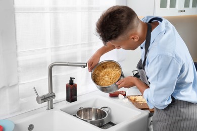 Photo of Man draining water from saucepan with pasta in messy kitchen