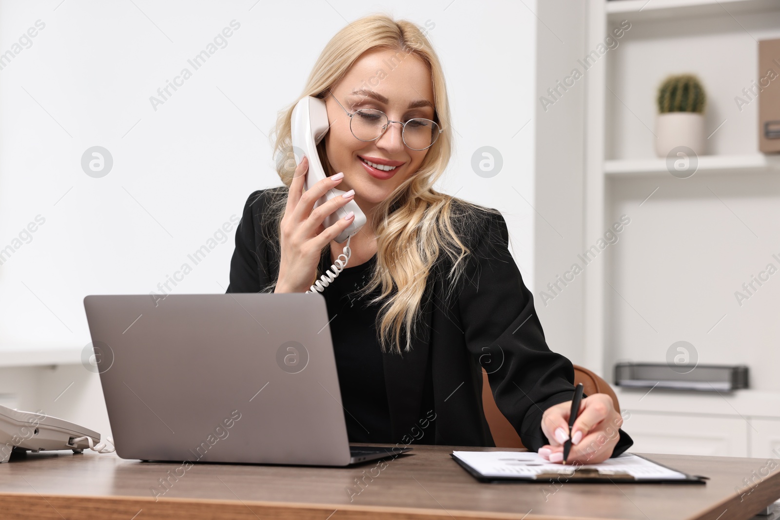 Photo of Happy secretary taking notes while talking on phone at table in office