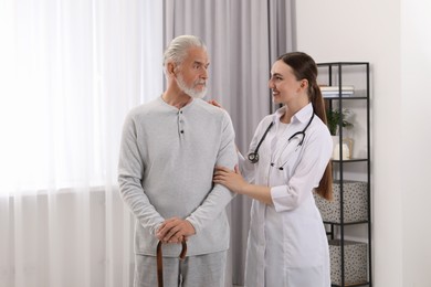 Photo of Young healthcare worker assisting senior man with walking cane indoors