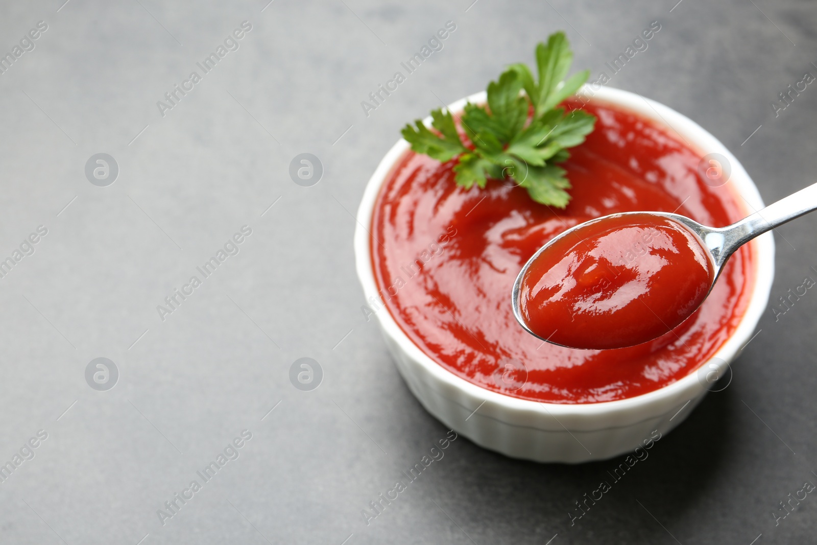 Photo of Taking delicious tomato ketchup with spoon from bowl at grey textured table, closeup. Space for text