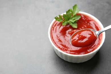 Photo of Taking delicious tomato ketchup with spoon from bowl at grey textured table, closeup. Space for text