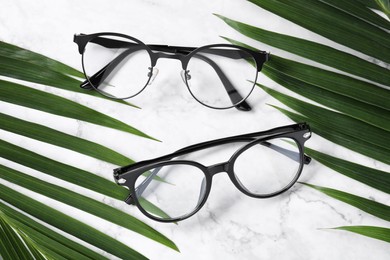 Glasses in stylish frames and tropical leaves on white marble table, flat lay