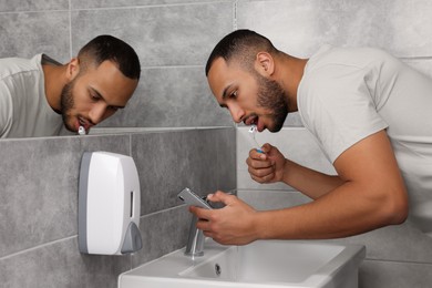 Young man using smartphone while brushing teeth in bathroom. Internet addiction