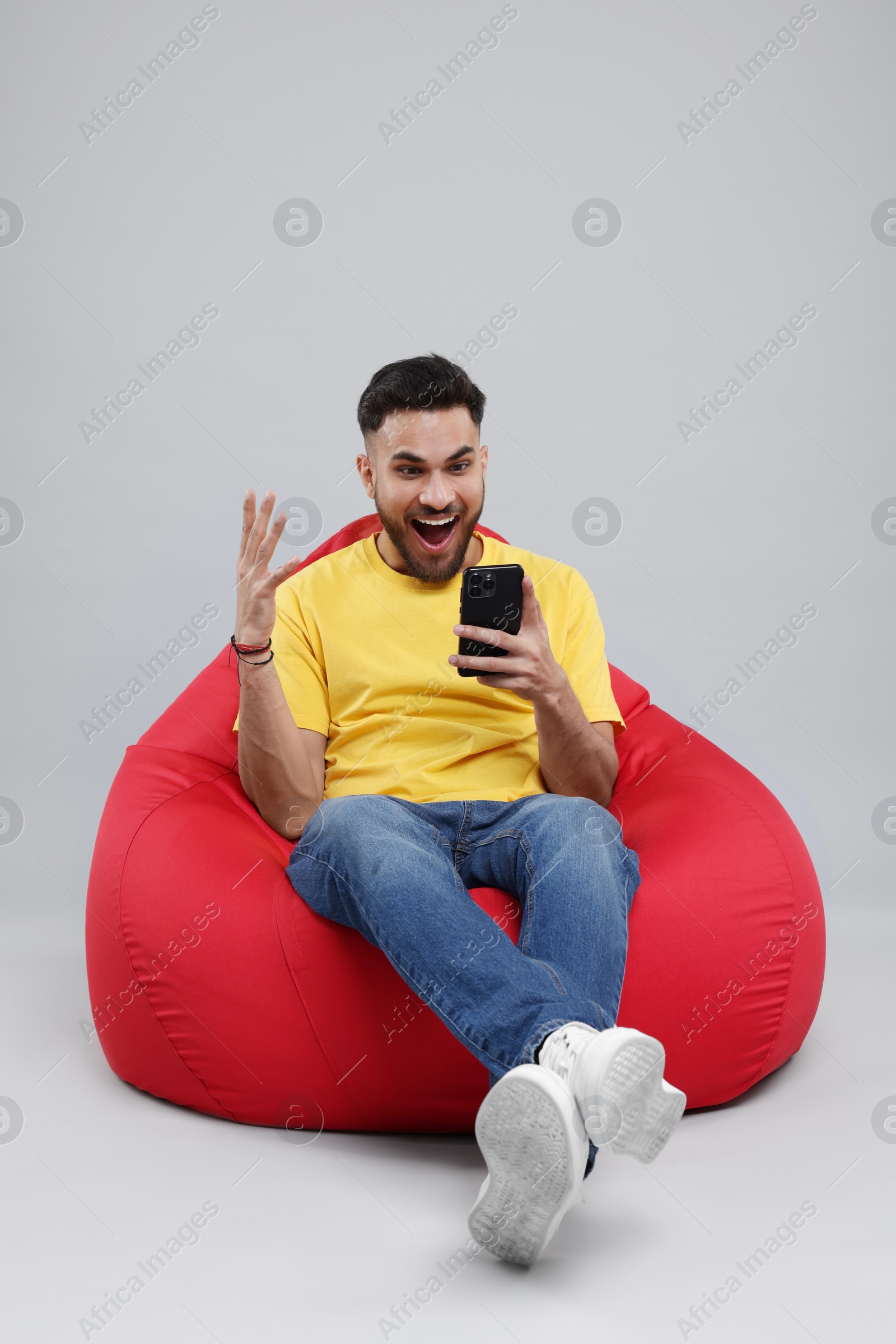 Photo of Emotional young man using smartphone on bean bag chair against grey background