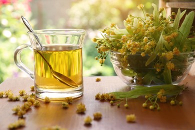 Photo of Glass cup of aromatic tea with spoon and linden blossoms on wooden table against blurred background