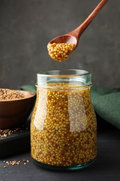 Photo of Taking whole grain mustard with spoon from jar on black wooden table