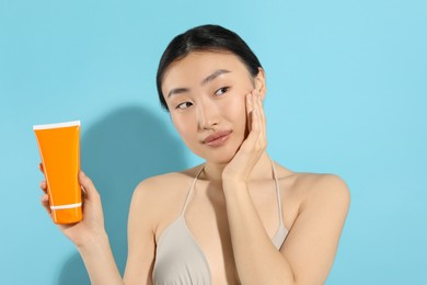 Beautiful young woman holding sun protection cream on light blue background