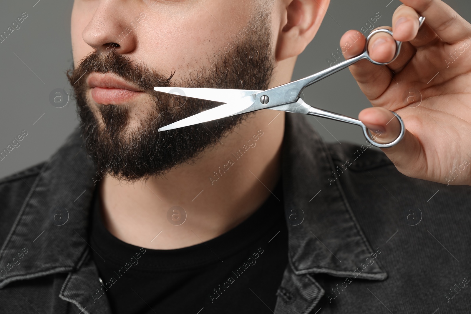 Photo of Handsome young man trimming beard with scissors on grey background, closeup