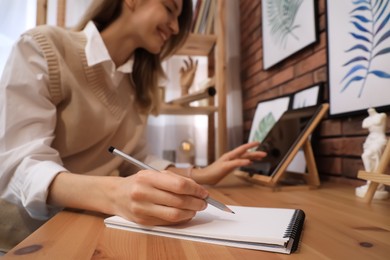 Photo of Young woman using tablet while drawing in sketchbook with pencil at wooden table indoors, focus on hand
