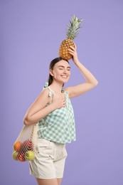 Photo of Woman with string bag of fresh fruits holding pineapple above her head on violet background