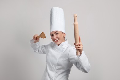 Photo of Happy professional confectioner in uniform holding wooden rolling pin and spatula on light grey background