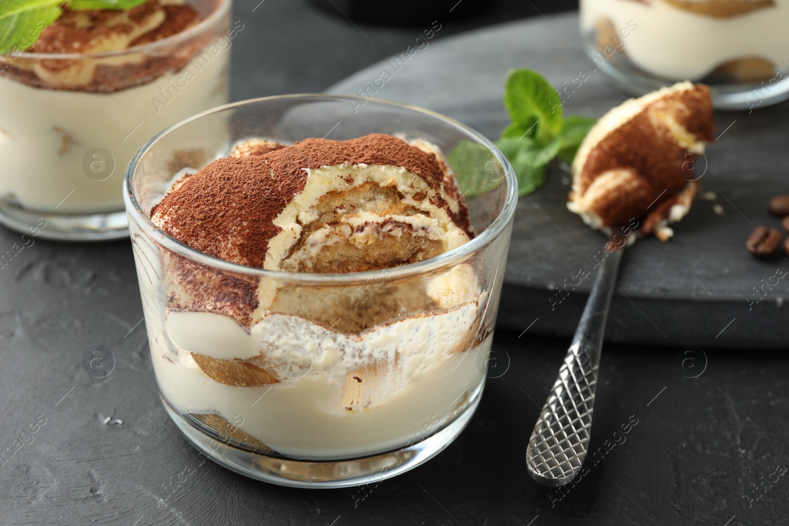 Photo of Delicious tiramisu in glasses and spoon on black table, closeup