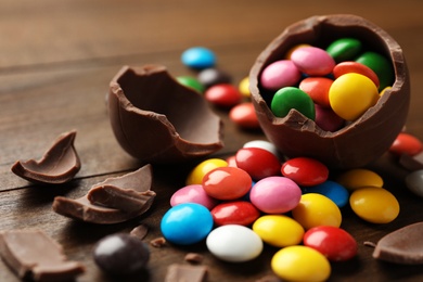 Photo of Broken chocolate egg and colorful candies on wooden table, closeup