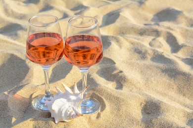 Glasses of tasty rose wine and seashell on sand, space for text