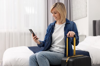 Smiling guest with smartphone and suitcase relaxing on bed in stylish hotel room