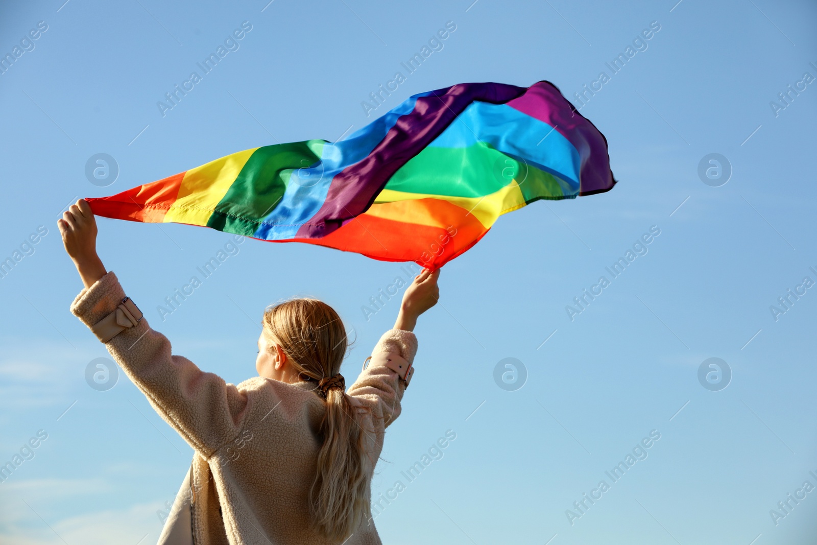 Photo of Woman holding bright LGBT flag against blue sky, back view