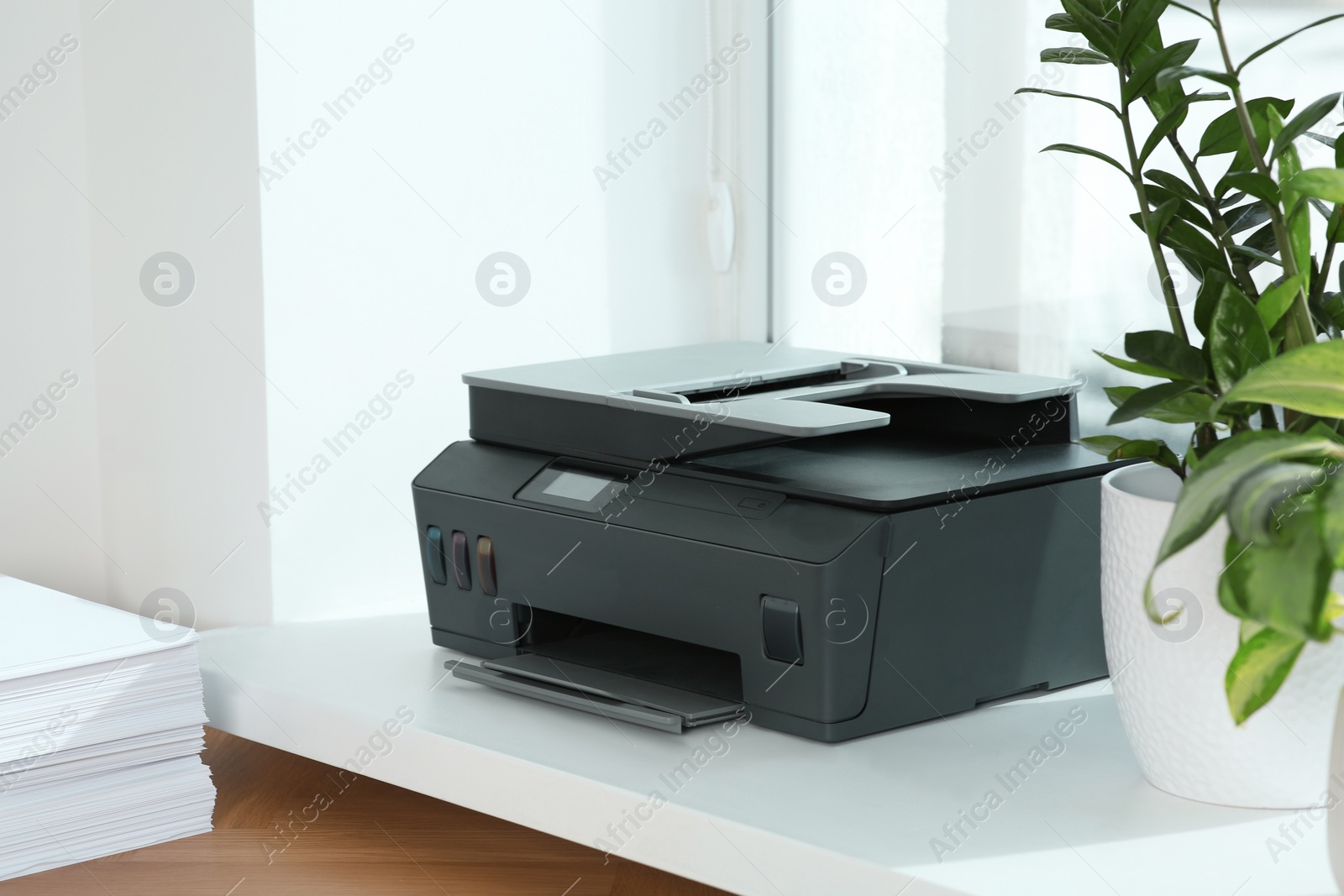 Photo of Stack of paper on wooden table near window sill with modern printer in office