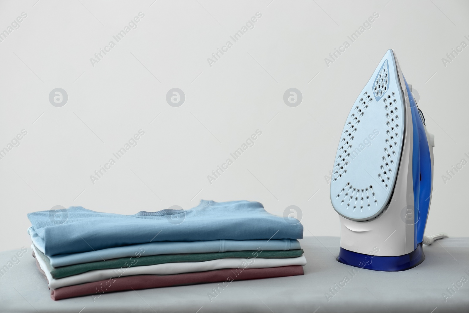 Photo of Modern iron and stack of clean clothes on board against white background