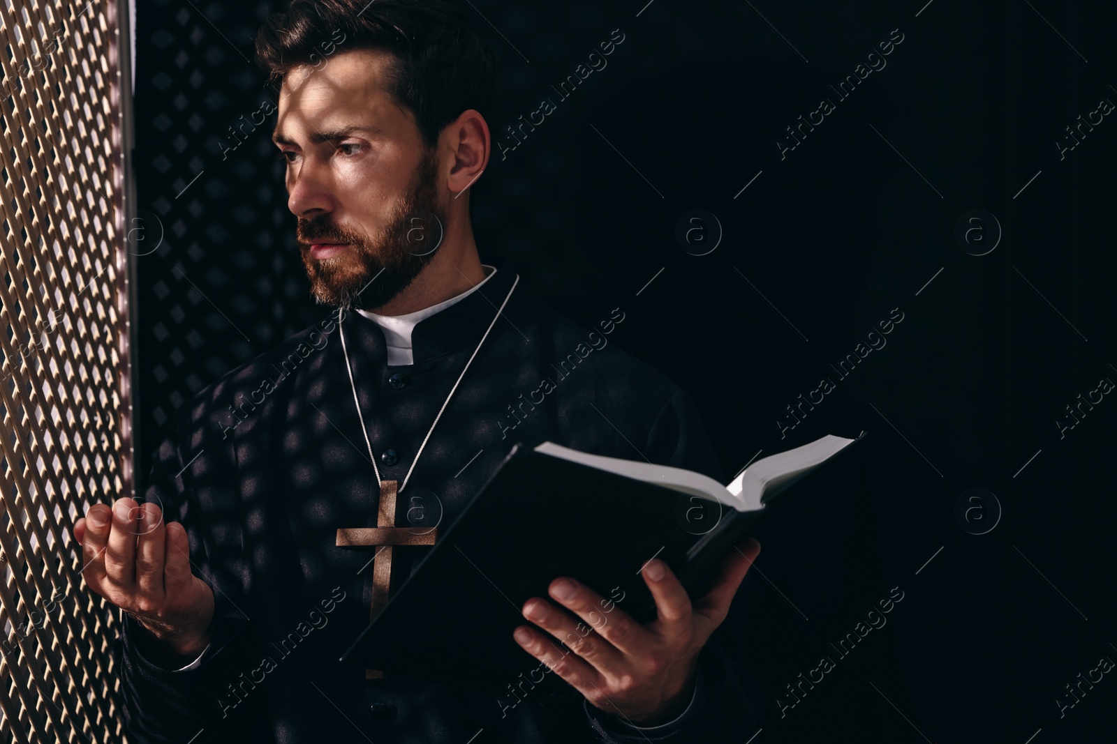 Photo of Catholic priest in cassock holding Bible and talking to parishioner in confessional