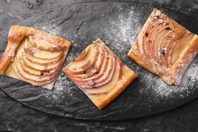 Photo of Pieces of delicious apple pie with powdered sugar on black table, top view