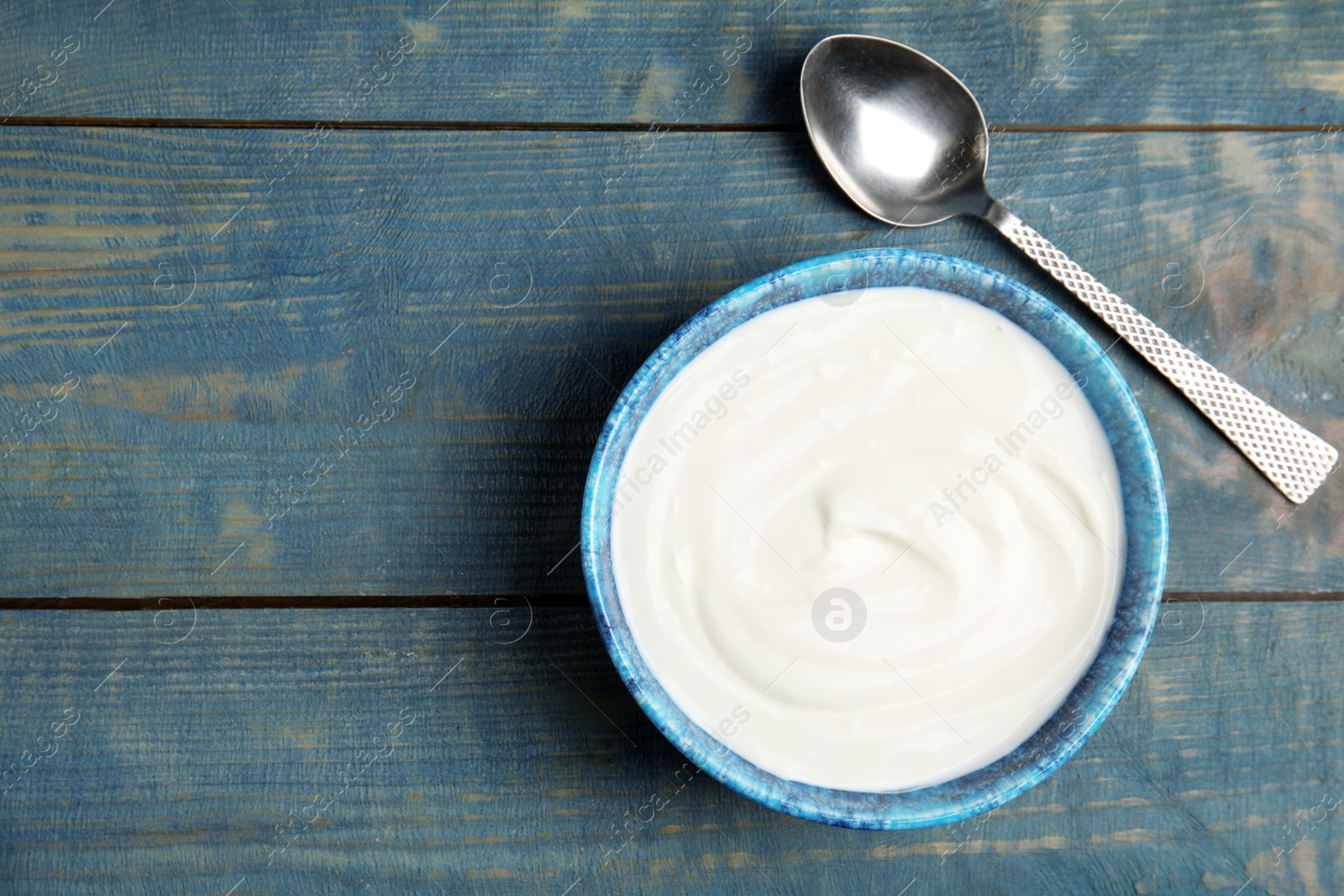 Photo of Bowl with creamy yogurt and spoon on blue wooden background, top view. Space for text