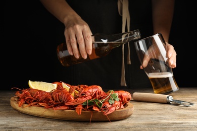 Photo of Delicious red boiled crayfishes on wooden table and woman pouring beer into glass on background, closeup