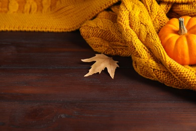 Photo of Orange knitted plaid with dry leaf and pumpkin on brown wooden table, closeup. Space for text