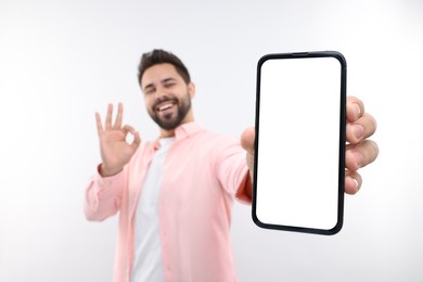 Photo of Young man showing smartphone in hand and OK gesture on white background, selective focus. Mockup for design