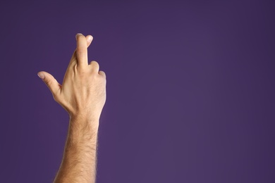 Photo of Man with crossed fingers and space for text on purple background, closeup. Superstition concept