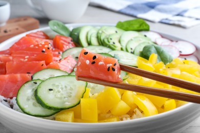 Wooden chopsticks with piece of salmon over delicious poke bowl on table, closeup