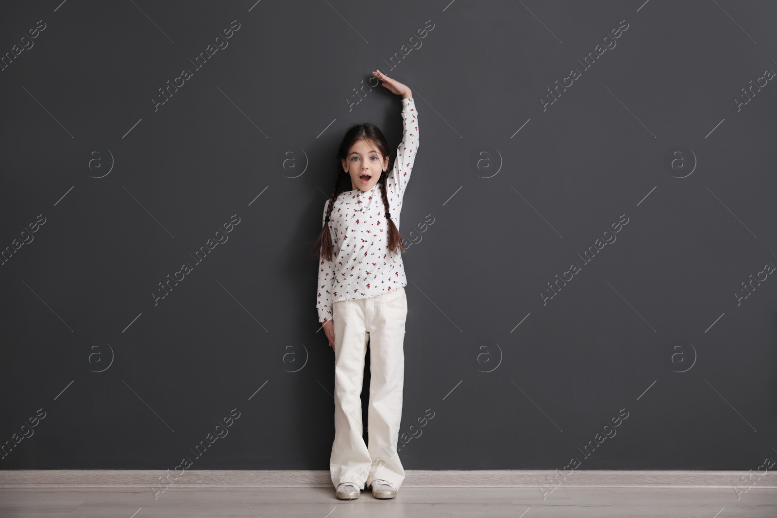 Photo of Emotional little child measuring her height near black wall indoors