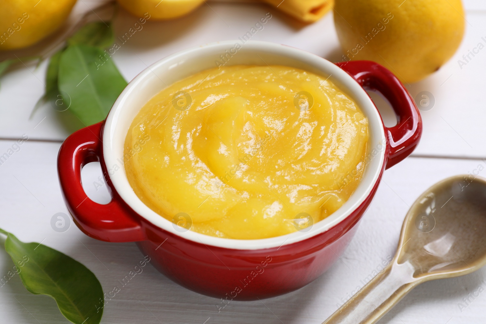 Photo of Delicious lemon curd in bowl, fresh citrus fruits and spoon on white table, closeup