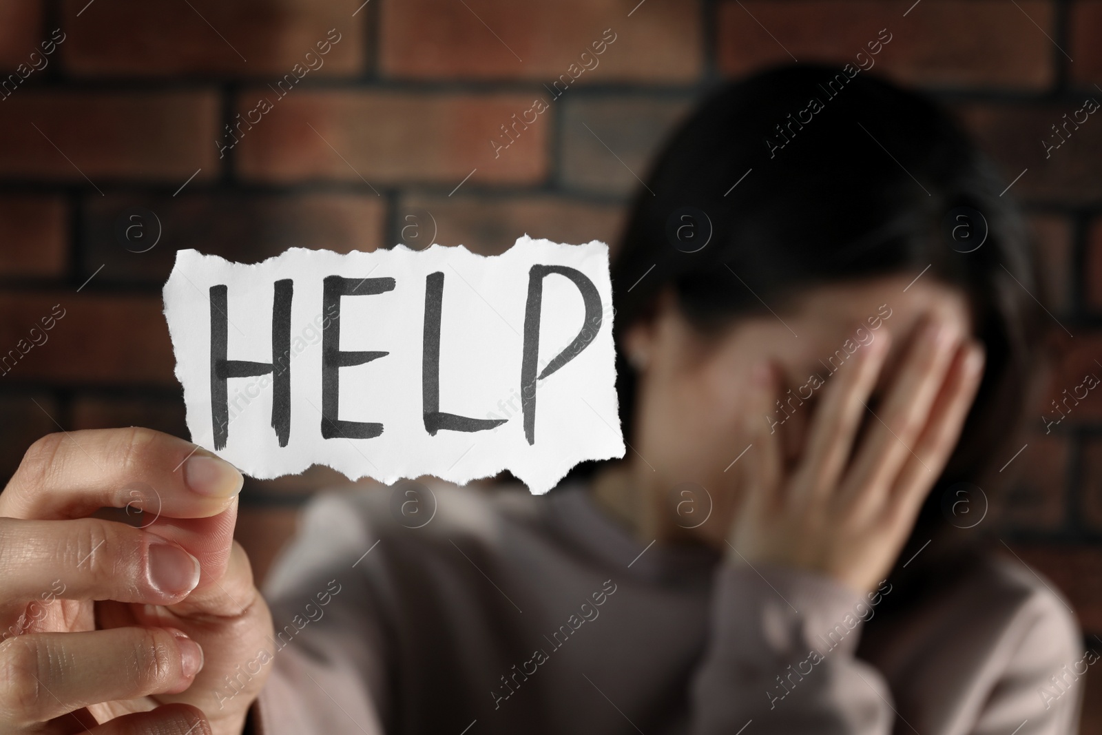 Photo of Domestic violence concept. Unhappy woman holding paper with written word Help near brick wall, focus on hand