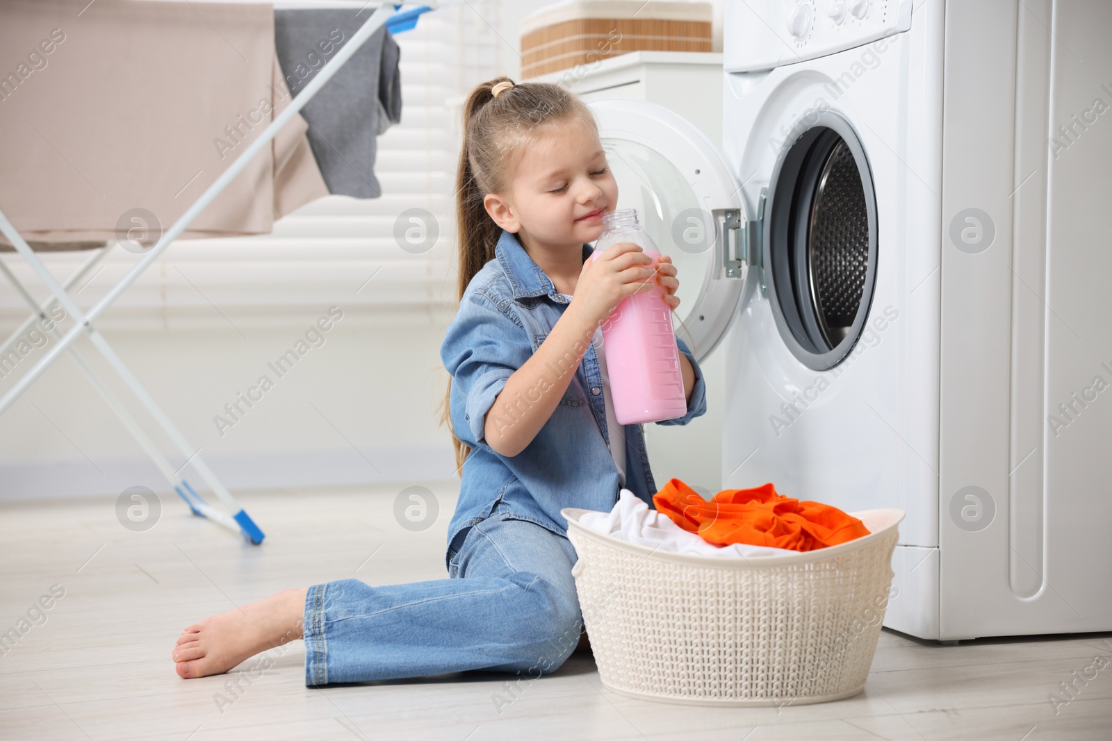 Photo of Little girl smelling fabric softener near washing machine and basket with dirty clothes in bathroom, space for text