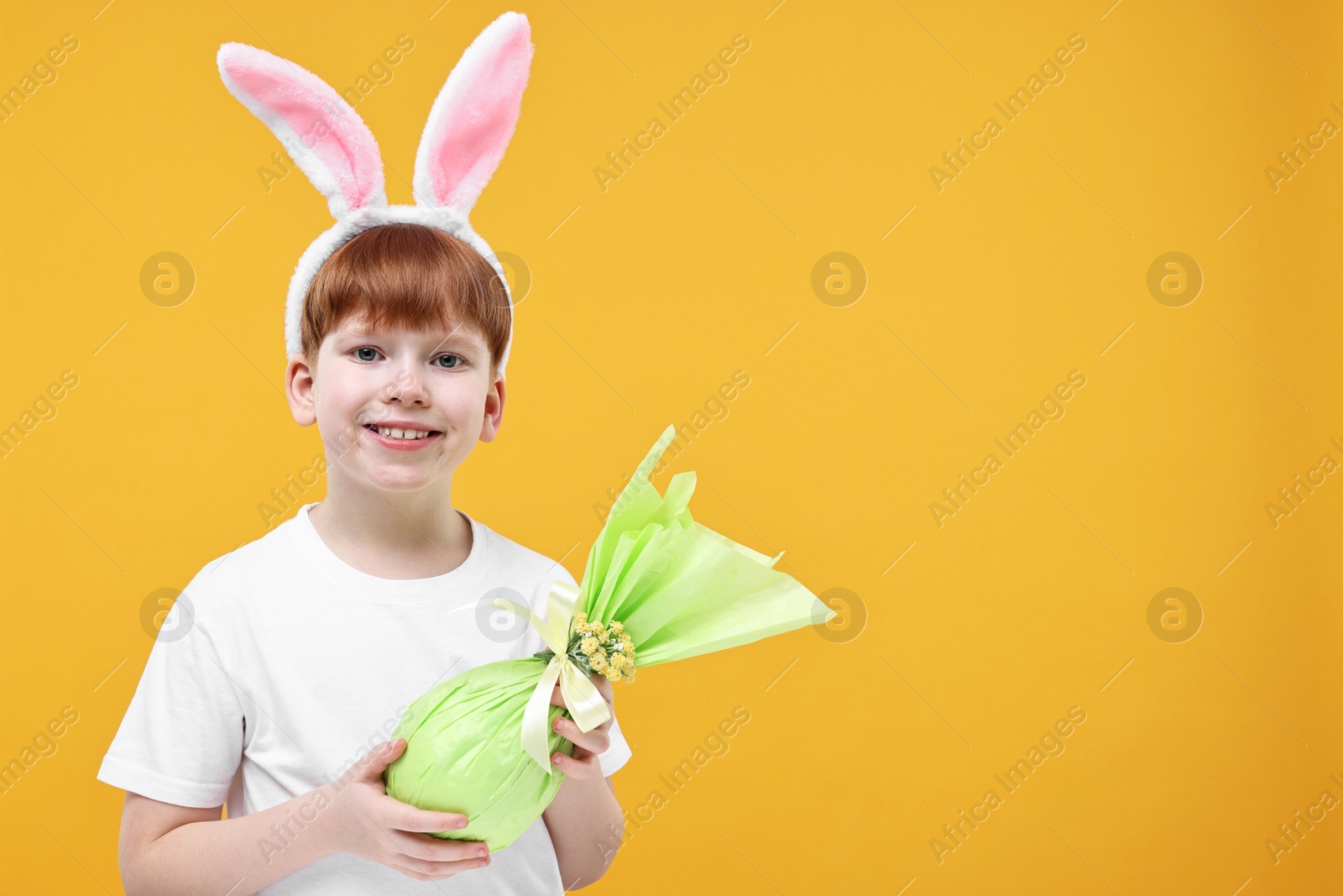 Photo of Easter celebration. Cute little boy with bunny ears and wrapped egg on orange background. Space for text
