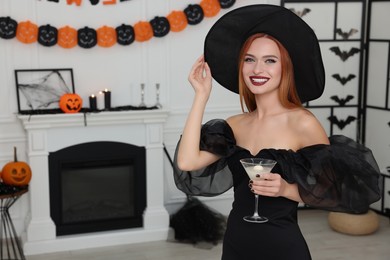 Photo of Happy young woman in scary witch costume with glass of cocktail indoors, space for text. Halloween celebration
