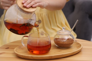 Woman pouring aromatic hot tea into glass cup at wooden table, closeup